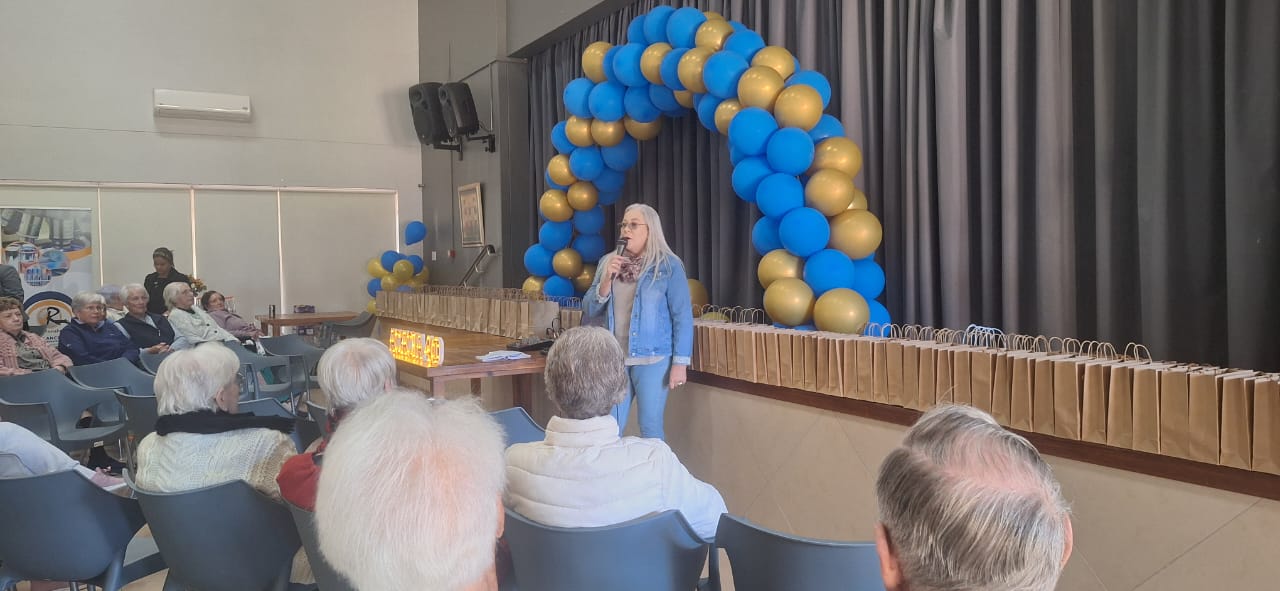 Karen speaking to a seated audience at a community event under a blue and gold balloon arch